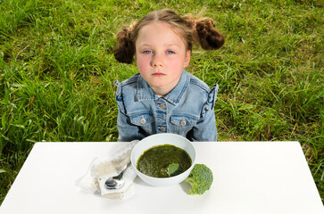 A dejected child sits behind a bowl of broccoli soup.