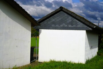 Zwei weiße Betonschuppen mit grauem Spitzdach auf Wiese auf dem Land vor Himmel mit grau-weißem dramatischen Wolkengebilde bei Sonne und Regen am Mittag im Spätsommer