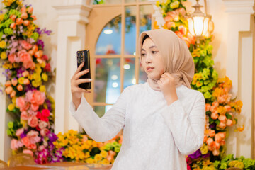 young woman hijab wearing elegant white muslim dress looking at mobile phone camera while adjusting hijab in luxury restaurant. for fashion, promotion, lifestyle content