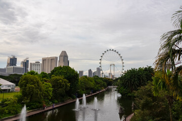 Naklejka premium Pathway winds along tranquil lake in Gardens by the Bay, Singapore. Trail is bordered by lush greenery and colorful flowers. Iconic Singapore Flyer Ferris wheel in background. Cityscape SouthEast Asia