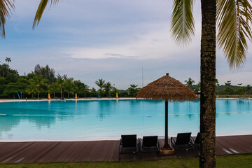 Lounge chairs and thatched umbrellas in tropical resort of luxury Treasure Bay hotel, Bintan Island, Indonesia. Exotic palm trees in front of large clear blue swimming pool. Recharge getaway. Vacation