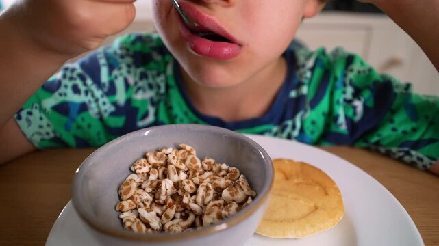 Young Boy eating cereal from a bowl, lifting the spoon to his mouth. A pancake sits on the plate, a simple and casual breakfast moment