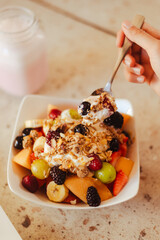 Manos sosteniendo una cuchara cargada con cereales, frutos rojos y cereales, debajo un bowl con un desayuno saludable
