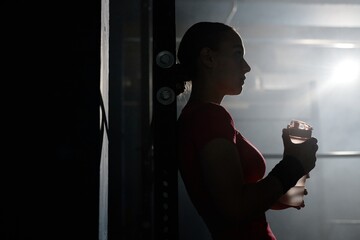 Silhouette of young woman standing against backlight holding bottle of sport drink while having break after intense workout in gym with low key lightning, copy space