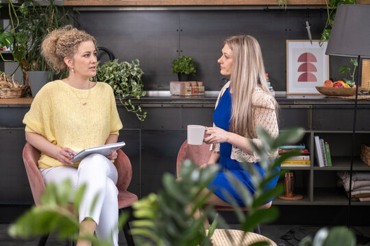 Conversation between two women in cozy living room, discussing ideas over coffee, surrounded by plants, teamwork concept