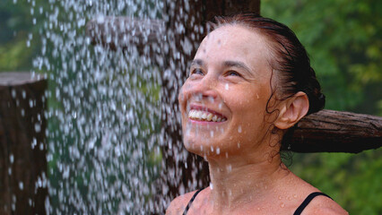 Woman smiles and takes shower in nature. Clip. Happy woman takes outdoor shower. Woman smiles while taking shower in jungle on summer day
