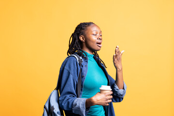 Sleepy teenager with rucksack yawning, feeling tired, drinking coffee to wake up on her way to university. Drowsy student using caffeinated beverage to gain energy, isolated over studio background