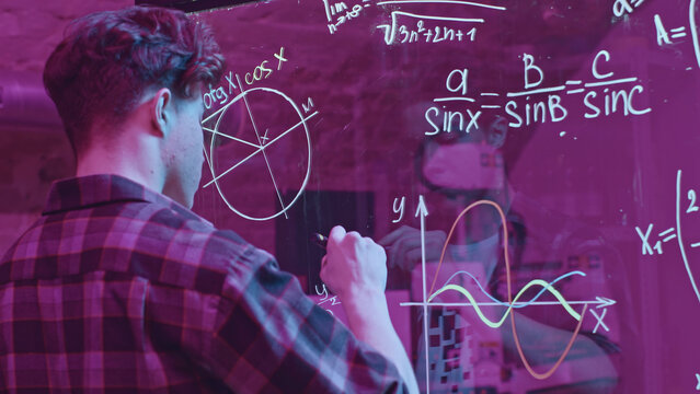 Young man solves mathematical equations on blackboard. Media. Young man solves complex equations on transparent board. Genius writes equations on blackboard in neon light - Powered by Adobe