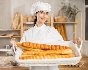 Near table in work room - space, baker girl in white uniform and hat stands with basket of baguettes. She shows products of baguettes made from different types of flour, assortment of French bakery