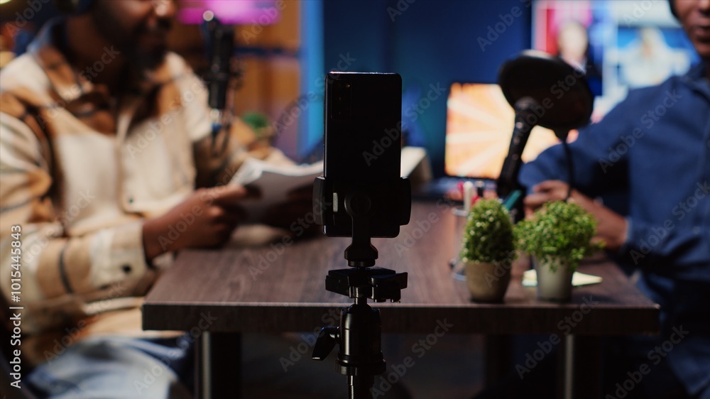 Poster focus on smartphone on tripod used by podcast host in blurry background to record guest telling inte
