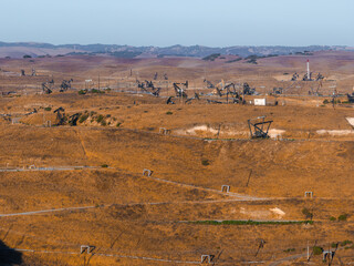 A vast desert landscape in California features numerous oil rigs amidst rolling hills. The scene includes power lines and small structures under a clear sky.