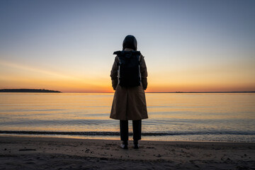 A girl in a coat with a backpack on the seashore at sunset