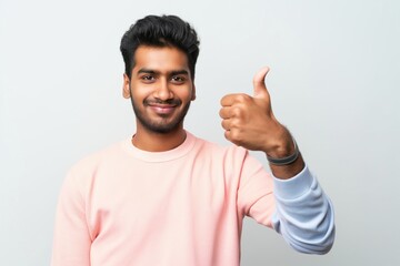 Fototapeta premium Young man stands against plain white background with thumbs up gesture. Wears casual outfit of pink shirt, blue jeans. Smile conveys sense of approval, positivity. Simple background puts focus on