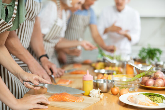 Pleased middle-aged female learner of culinary classes processing piece of salmon standing around kitchen table together with others