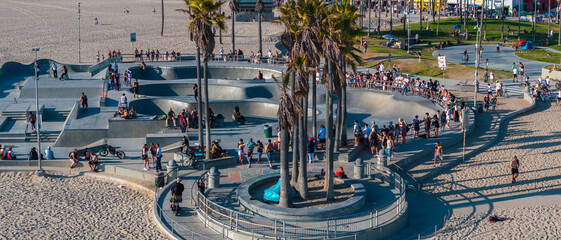 Venice Beach Skatepark in Los Angeles features skateboarders on ramps, tall palm trees, sandy beach, ocean, and a crowd of onlookers. © Aerial Film Studio
