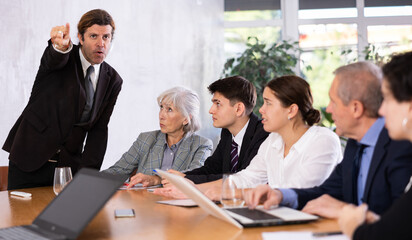 Fototapeta premium Furious male boss yelling at office colleagues of different ages taking notes at table with laptops
