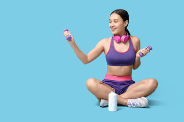 Young woman in sportswear and with dumbbells sitting against blue background