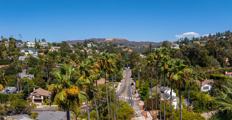 The image captures the Hollywood district in Los Angeles, featuring the Hollywood Hills and Sign. A tree lined street with palm trees and houses is visible. © Aerial Film Studio