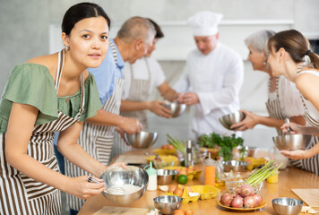 Pleased middle-aged woman learner of culinary classes making sauce in metal basing mixing it with metal whisk