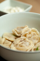 Steaming bowl of dumplings seasoned with black pepper and dill, ready to eat. Perfect for lunch or dinner, served with sour cream on a white plate