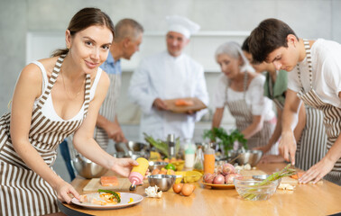 Naklejka premium Positive young woman attendee of cooking course decorating meal made of salmon fish together with others