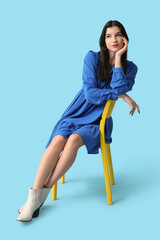 Beautiful young woman in stylish blue dress on chair against color background
