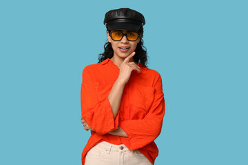 Stylish African-American woman wearing sunglasses and hat on blue background