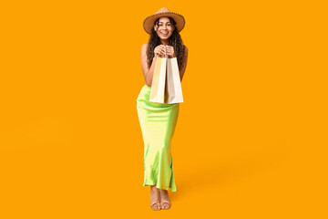 Stylish African-American woman in dress and hat holding shopping bags on yellow background