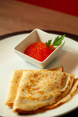 Thin golden pancakes with a side of red caviar and fresh parsley are served on a white plate. This simple dish is a classic russian appetizer