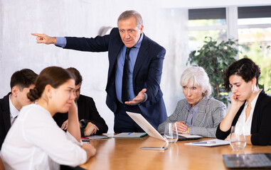 Fototapeta premium Angry elderly male boss scolding office workers of different ages using notebooks at table in office