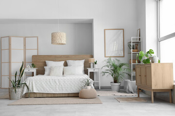 Beautiful interior of light bedroom with wooden chest of drawers, window, houseplants and bed