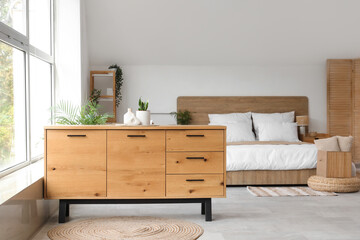 Stylish interior of light bedroom with wooden chest of drawers, houseplant and bed