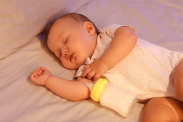 Cute little baby with bottle of milk sleeping on bed at night, closeup