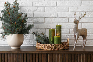 Vase with Christmas branches, burning candles and toy reindeer on commode near white brick wall