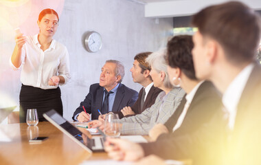 Colleagues of different ages using laptops and watching presentation while young female turning on presentation in office