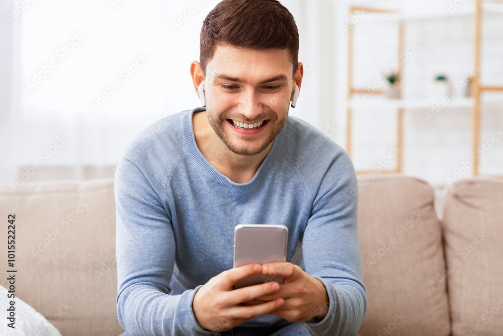 Poster Mobile User. Joyful Man In Wirelss Earphones Using Smatphone Playing Games Sitting On Sofa Indoor.