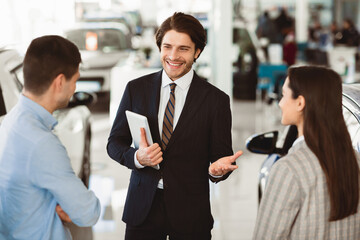 Professional Car Seller Talking With Young Couple Clients Selling Auto Working In Dealership Center. Selective Focus