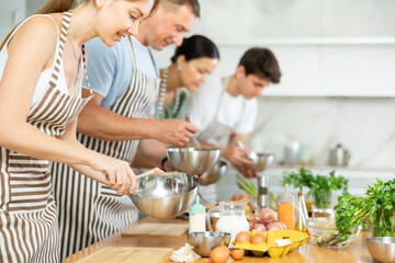 Positive interested young woman attending group culinary classes, standing by table with ingredients and utensils, learning culinary skills from professional chef
