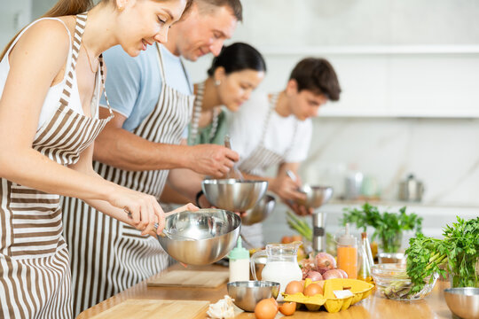 Positive interested young woman attending group culinary classes, standing by table with ingredients and utensils, learning culinary skills from professional chef