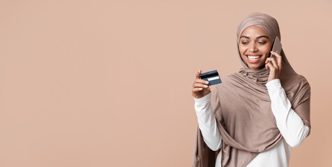 Fototapeta premium Customer service concept. Young black muslim woman in hijab holding credit card and talking on cellphone with bank manager, yellow background