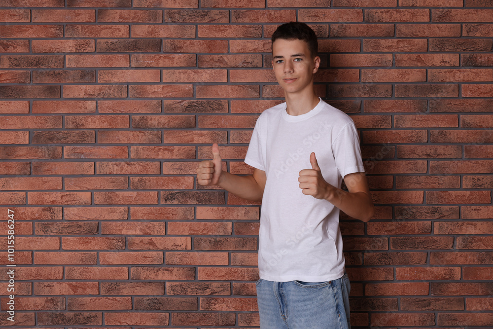 Poster Teenage boy wearing white t-shirt and showing thumbs up near brick wall, space for text