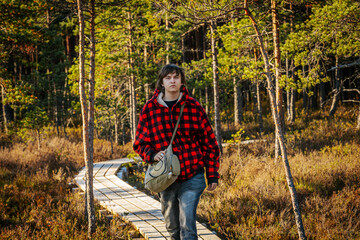 A young man wearing a red and black plaid jacket walks on a wooden boardwalk through a dense forest...