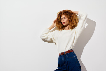 Fashion portrait of a young woman with curly hair wearing a stylish white sweater and high waisted jeans against a minimalist white background