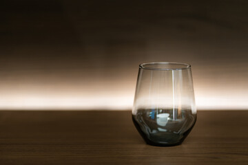 A single, empty glass cup sits on a dark wooden table. The glass has a curved bottom and a wide rim. The background is a gradient of dark brown to a lighter brown, with a thin line of light at the bot