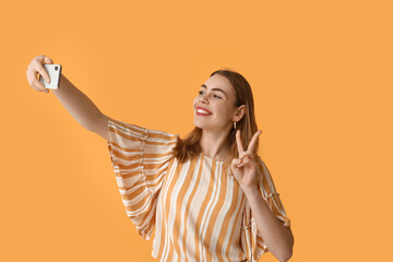 Fototapeta premium Pretty young woman showing peace gesture while taking selfie on orange background