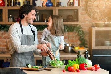 Happy father and little daughter cooking vegetable salad in kitchen, adding seasoning and laughing together while preparing lunch at home