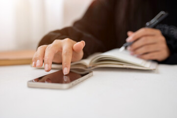 A close-up of a woman writing in a book and using her smartphone while sitting at a table indoors.