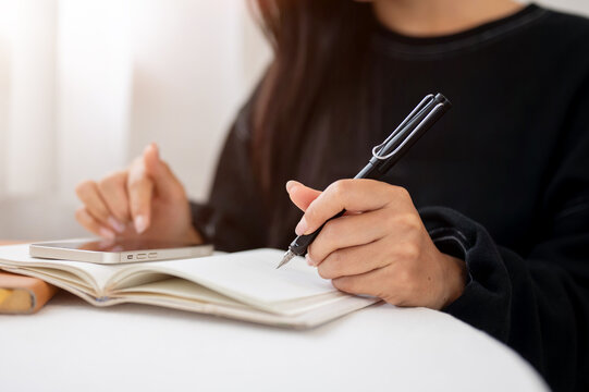 A close-up of a woman writing in a book and using her smartphone while sitting at a table indoors.