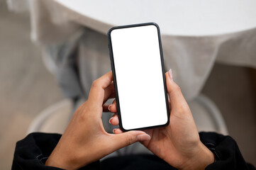 A woman holding a smartphone with a white screen mockup while sitting at an indoors table.