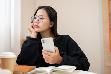 A thoughtful Asian woman gazes out the window while sitting at an indoor table, deep in thought.